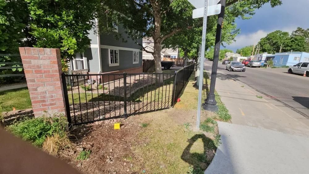 Black metal fence in front of a gray house, next to a brick pillar and street.