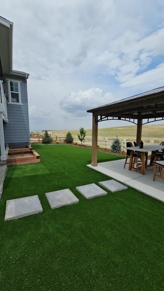 Backyard with green lawn, stone pavers, a patio with a pergola, and a light blue house on a cloudy day.