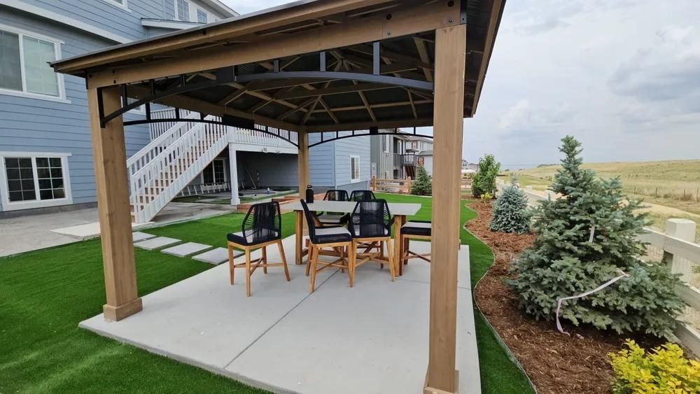 Gazebo with table and chairs on a concrete patio, green turf lawn, and landscaping in a backyard setting.