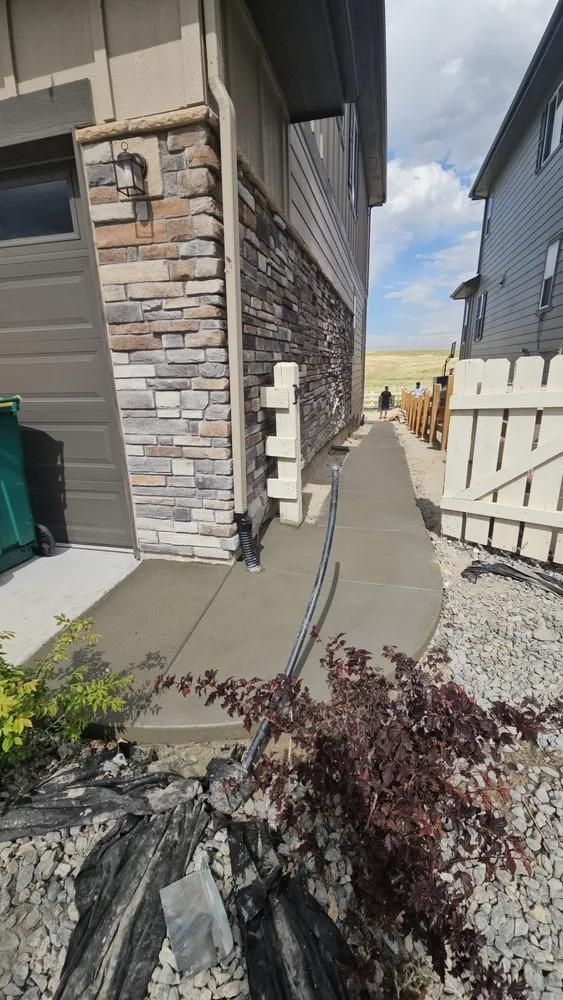 New concrete walkway beside a house with stone veneer, leading towards a wooden fence and grassy hill.