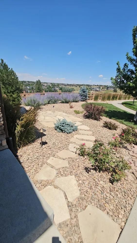 Stone path through a xeriscape garden with purple flowers, overlooking a distant landscape under a blue sky.