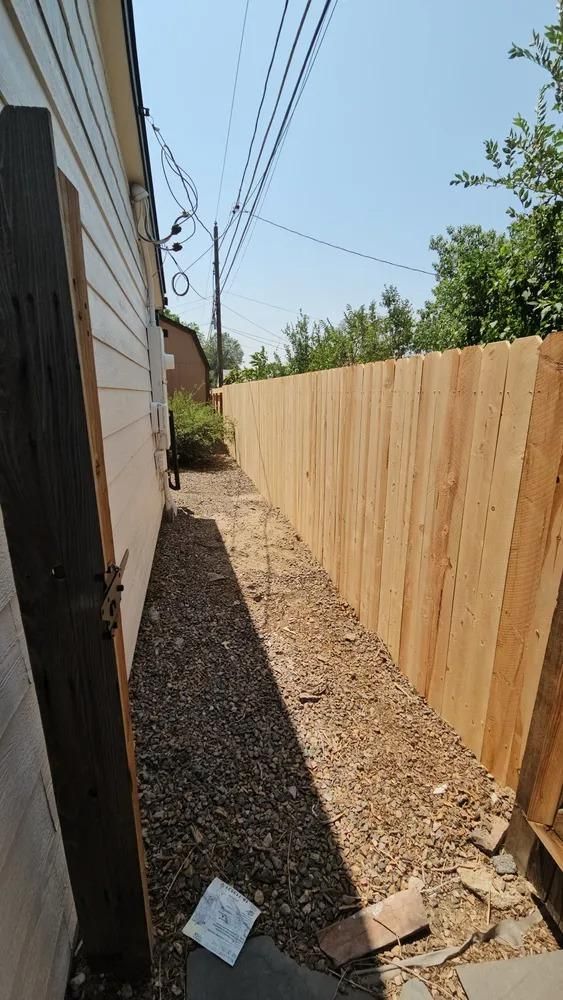 Gravel path between a wooden fence and a house with light siding on a sunny day. 