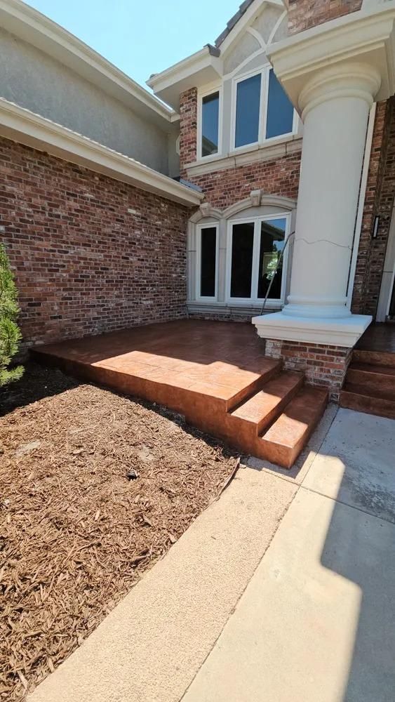 Brick-faced house entrance with steps and a large porch, bordered by mulch and a sidewalk.