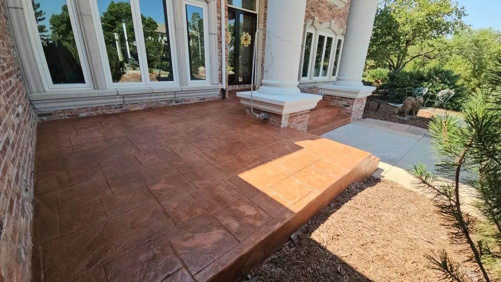 Brown stamped concrete porch with brick wall, white columns, and landscaping.