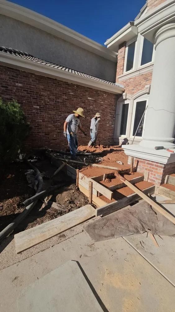 Two construction workers building steps in front of a brick house with a column, under a clear, blue sky.