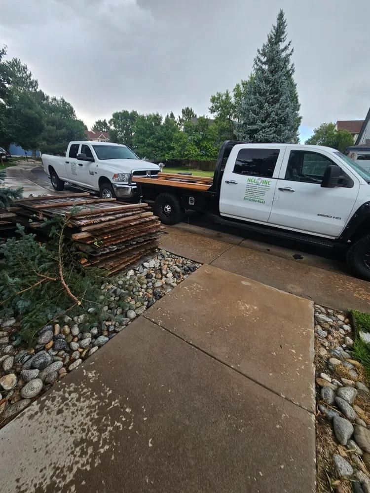 Two white trucks, one pulling wood planks, parked on a driveway next to a walkway with rocks.