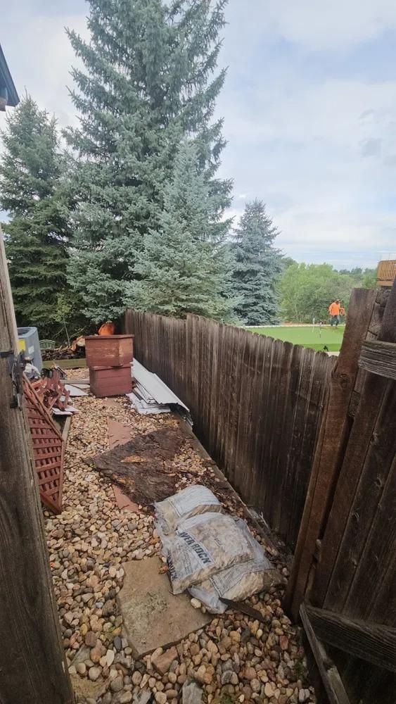 Backyard with wooden fence, gravel, debris, and blue-green evergreen trees under cloudy sky.