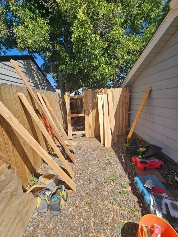 Wooden fence with a gate, tools, and lumber on gravel. Bright sunlight.