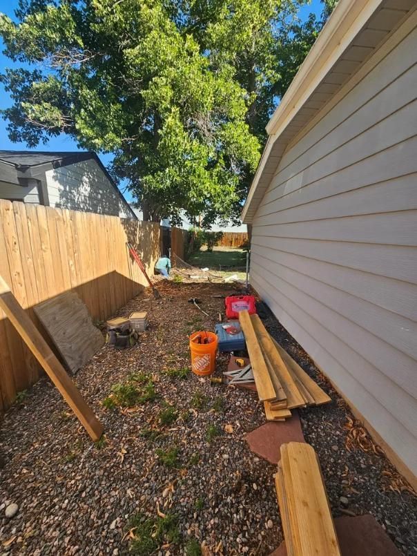 Backyard construction site; wooden fence, new siding on a white building, tools, and lumber on the ground.