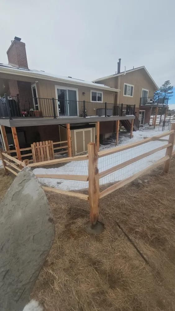 Wooden split-rail fence with chicken wire in front of a house with a deck. 