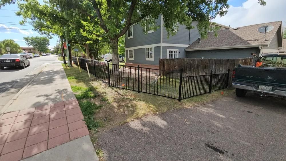 Gray two-story house with black fence next to a sidewalk and a street with parked cars.