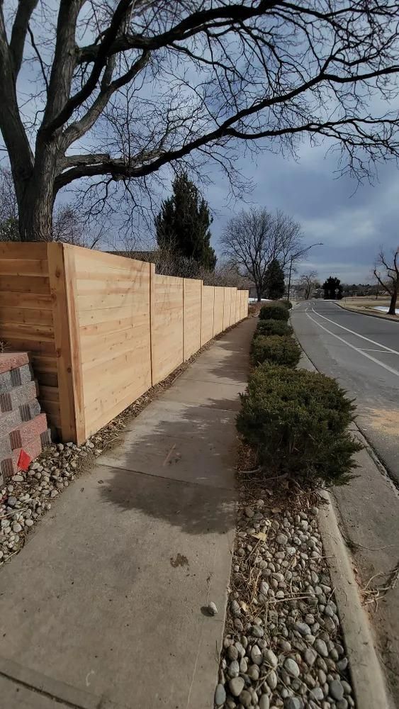 Sidewalk along a road with a wooden fence, bushes, and a tree under a cloudy sky.