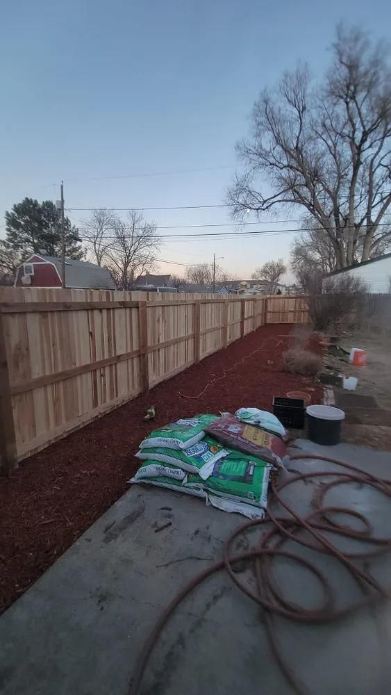 Backyard with new wooden fence, reddish mulch, bags of soil, and a hose on concrete.