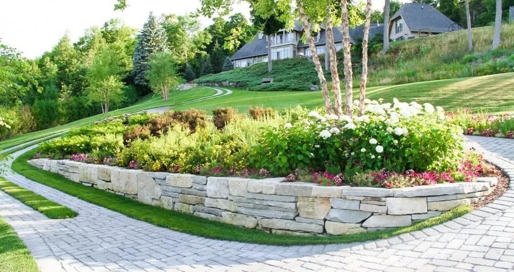 Stone retaining wall with lush garden, curving driveway, and house on a hill.