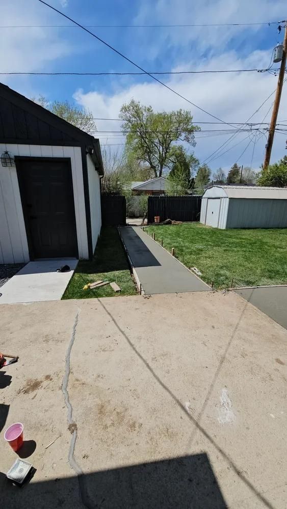 Concrete pathway leading to a backyard between a garage and fence under a blue sky.