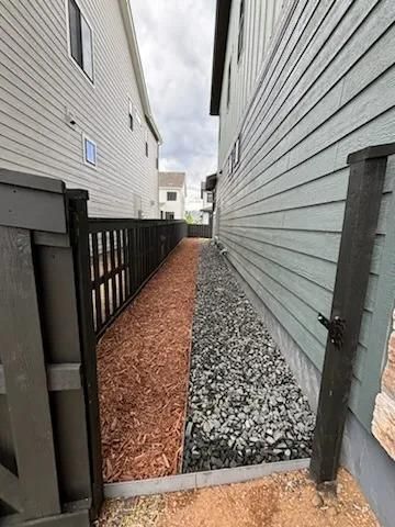 Narrow walkway between two houses, one side with wood chips, other with gravel, fenced.
