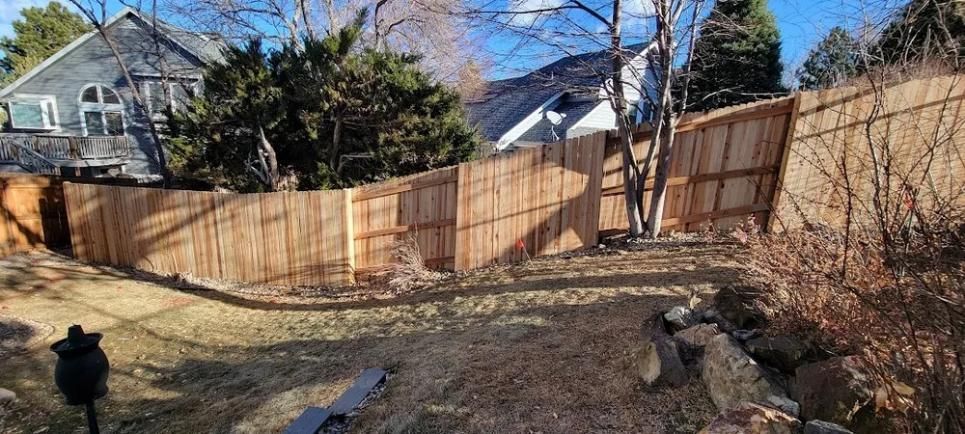 Wooden fence surrounds a backyard with a house, trees, and dry grass.