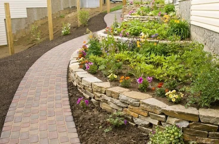 Brick pathway winds past tiered stone-walled garden beds with colorful flowers.