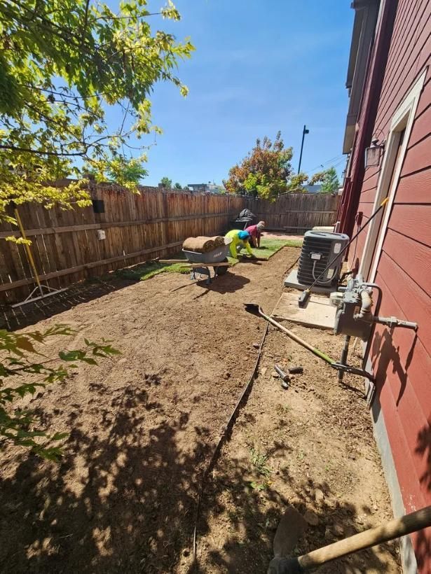 Yard work in progress: Two people near wheelbarrow, brown dirt, wooden fence, red house, and a sunny day.