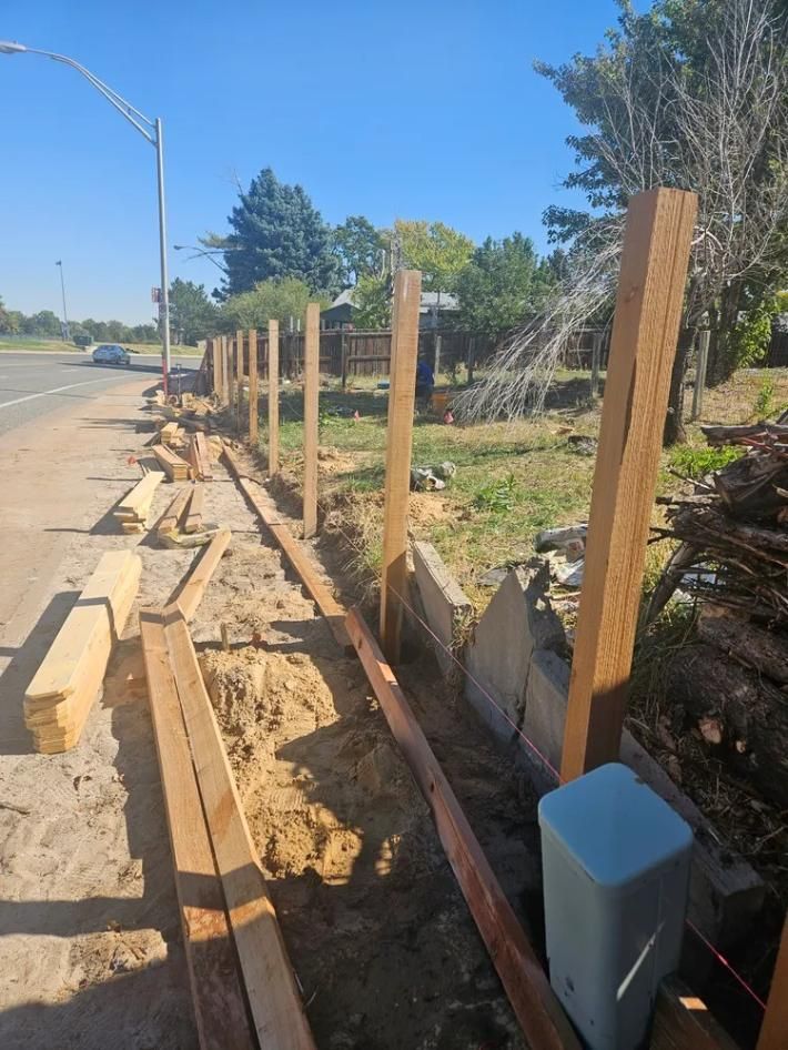 Wooden fence construction in progress next to a road, with posts in the ground and lumber laid out.