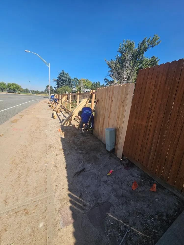 Construction workers building a wooden fence along a sidewalk, with a road and clear blue sky.
