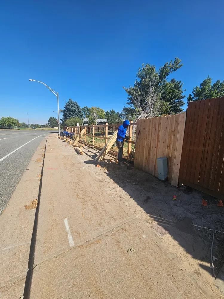 Workers constructing a wooden fence along a sidewalk next to a road on a sunny day.