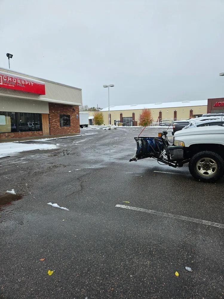 A snowplow truck clearing a wet parking lot next to a brick building under a cloudy sky.