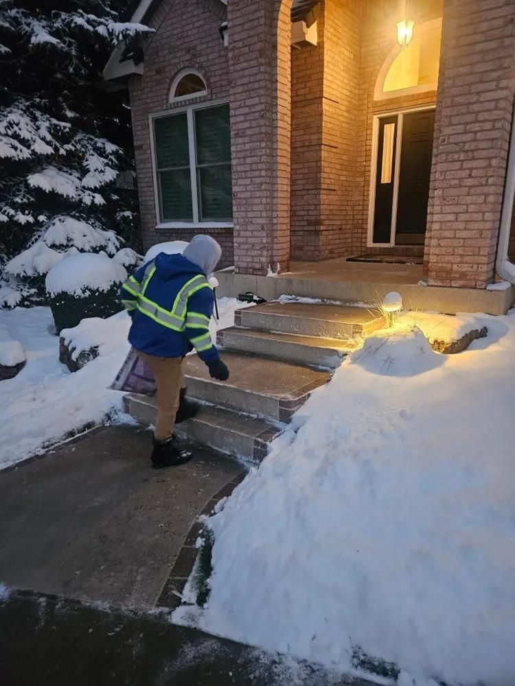 Person shoveling snow from steps of a brick house at dusk, wearing a reflective jacket.