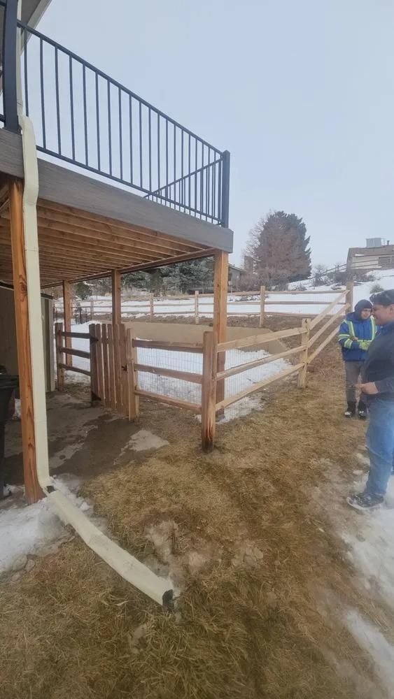 A wooden fence with a gate extends from under a deck, with two people standing near it in a snowy setting.