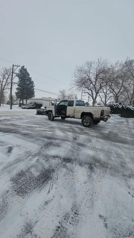 A white pickup truck with a snowplow is parked in a snowy parking lot.