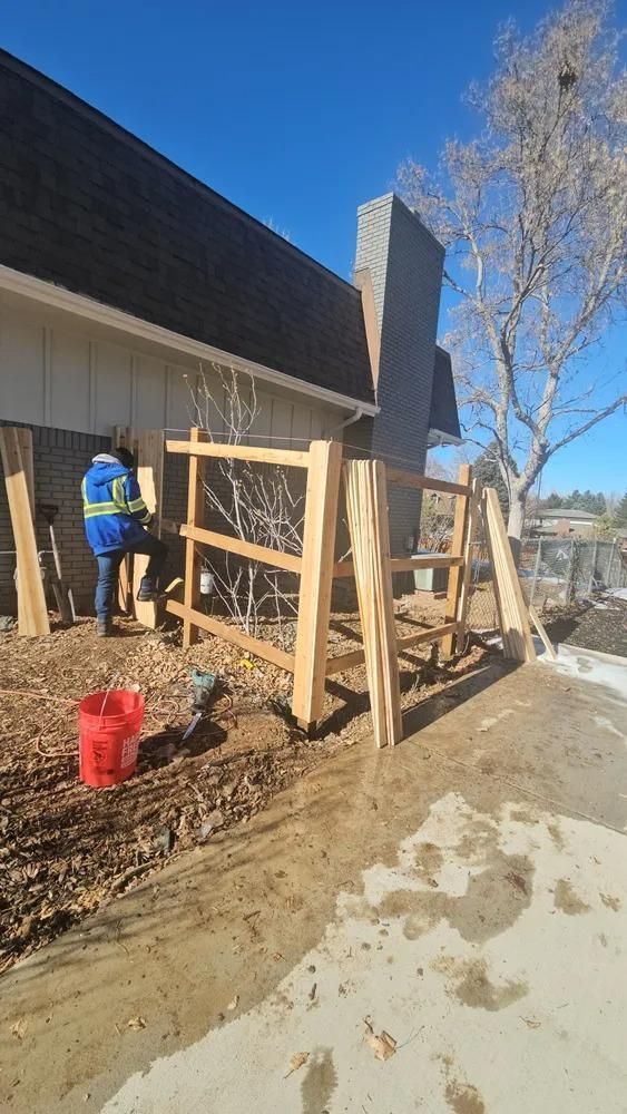 Person building a wooden fence next to a brick chimney and house. 