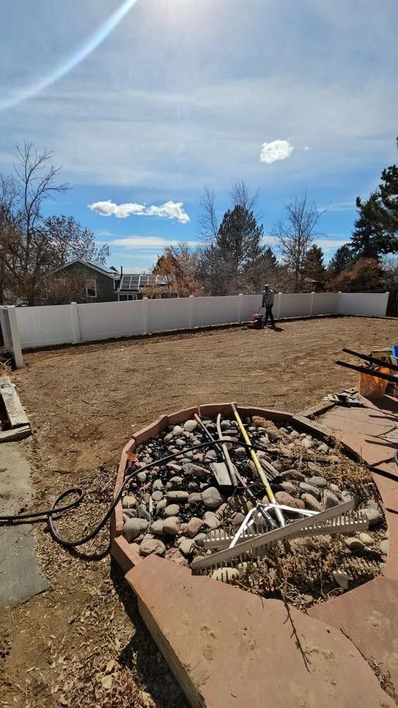 Backyard with a gravel surface, white fence, and person using a lawn tool under a bright sky.