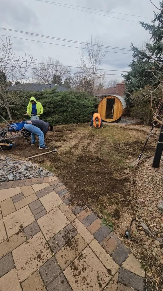 People working on a yard, preparing the ground with a sauna in the background. 