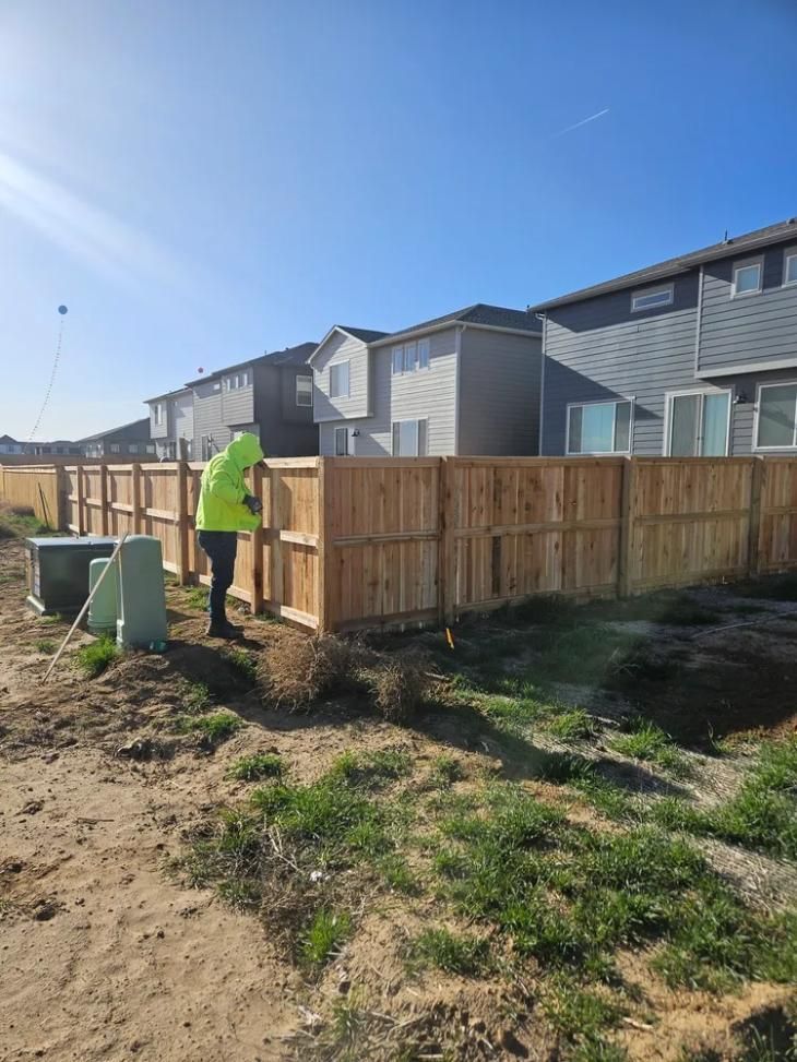 Person in safety vest near a newly built wooden fence, houses in the background, sunny day.