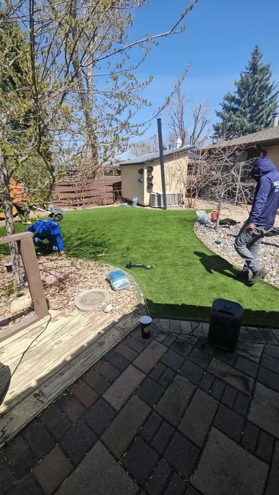Workers installing artificial turf in a backyard with a deck, trees, and buildings.