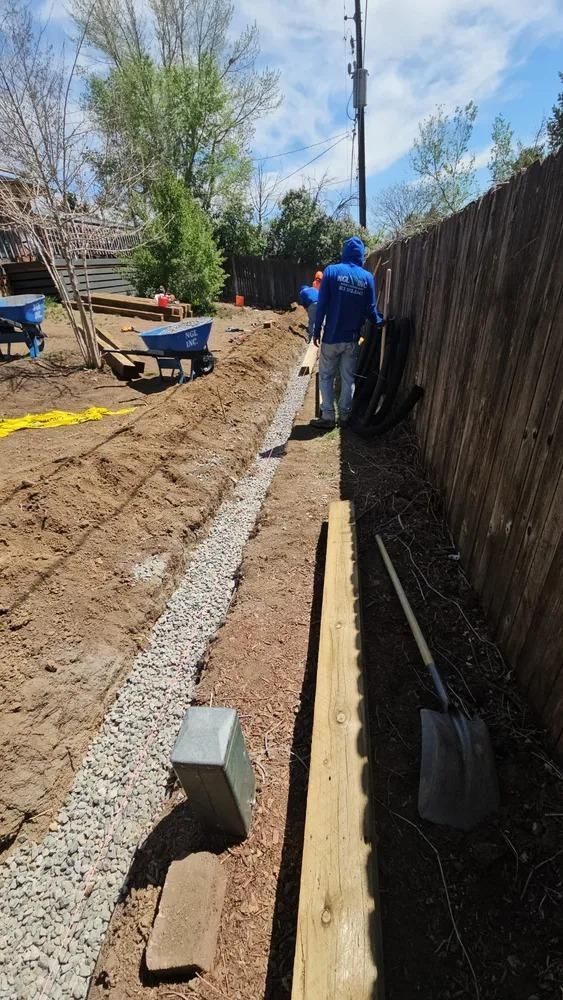 Person working in a trench, laying gravel near a wooden fence, outdoors.