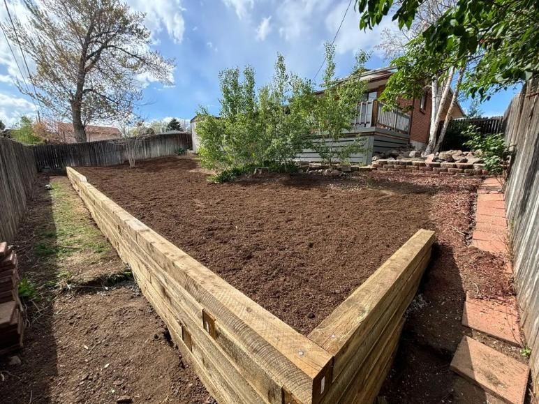 Wooden retaining wall in backyard with freshly tilled soil, surrounded by fences and a house.