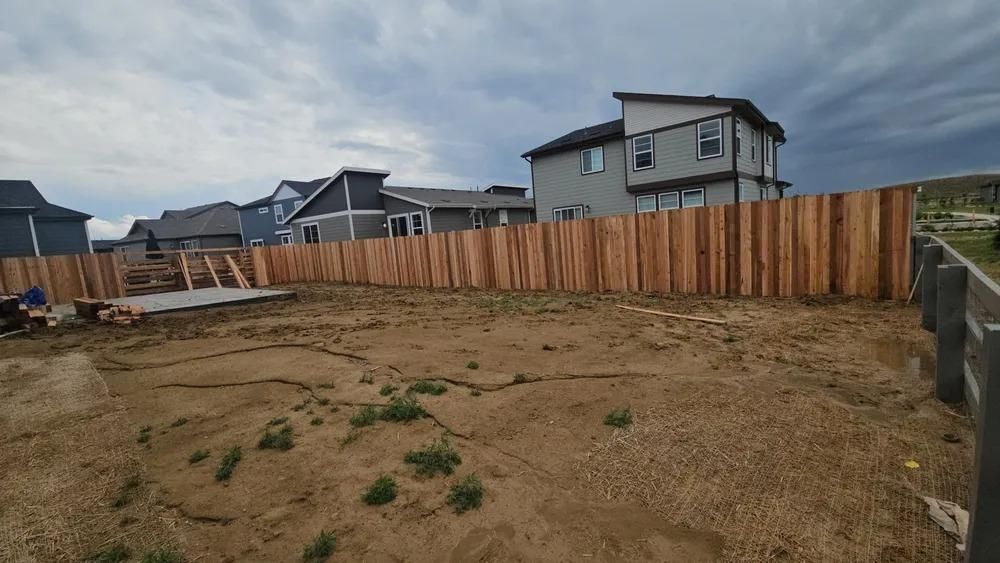Wooden fence surrounding a dirt yard, houses in the background, cloudy sky.