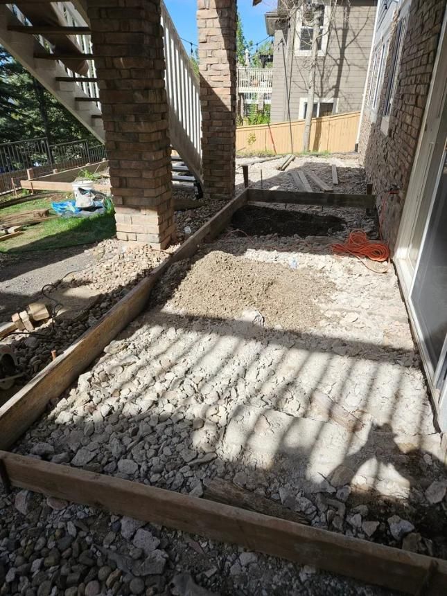 Construction site: framed ground with gravel and dirt, under a building with columns and stairs.