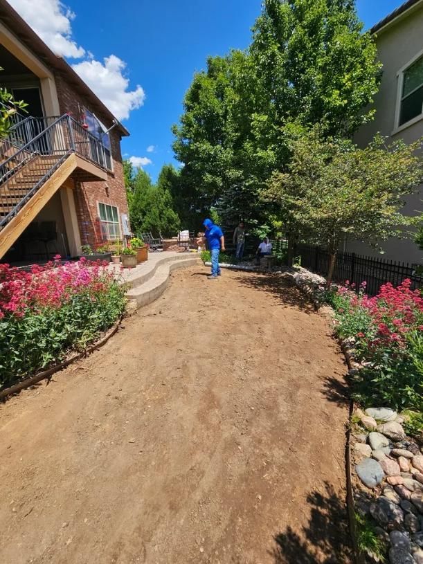 A person in blue stands on a dirt path in a garden, flowers on either side. 