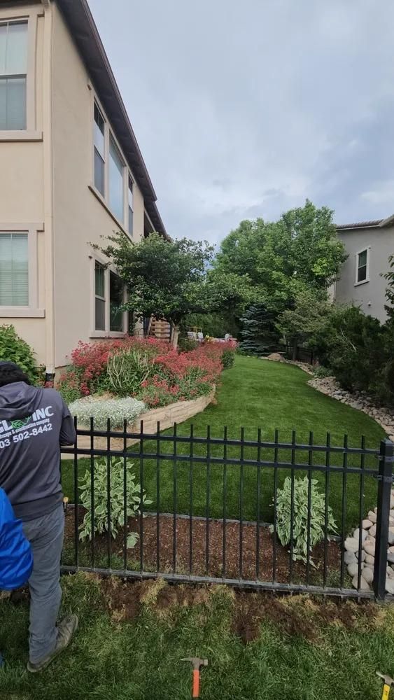 Man next to a house with a black fence, flowerbeds, and green grass. 