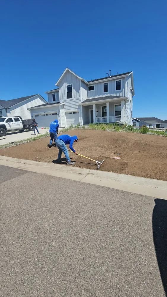 People raking a lawn in front of a light blue two-story house on a sunny day.