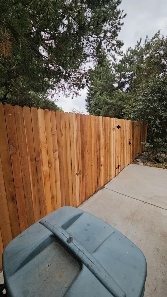 Wooden fence along a concrete patio, with a blue trash can in the foreground and trees in the background.