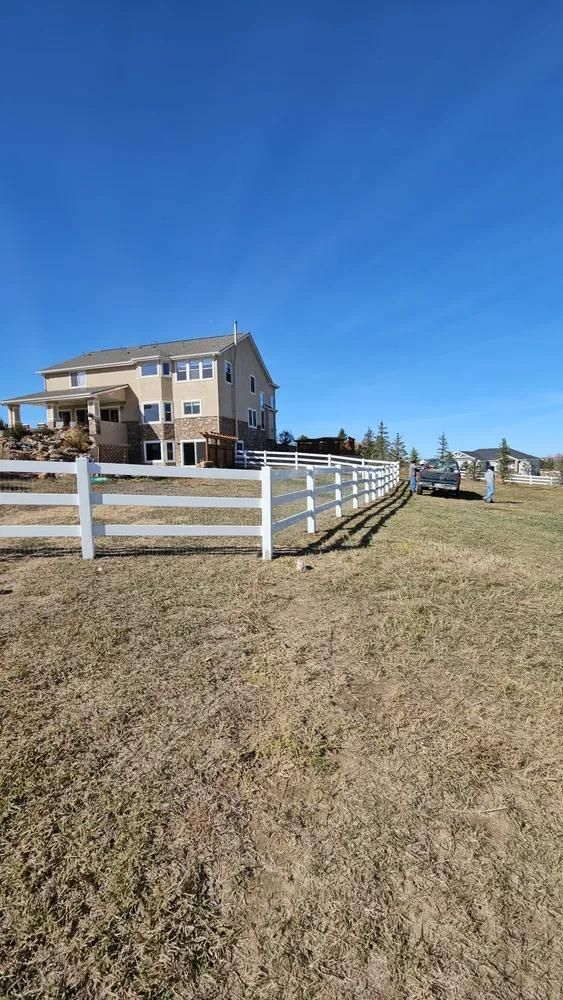 White fence in front of a two-story beige house under a clear blue sky.