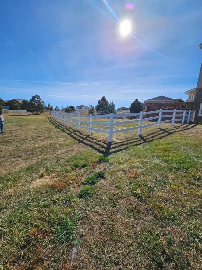 White three-rail fence in a grassy yard under a bright blue sky. 