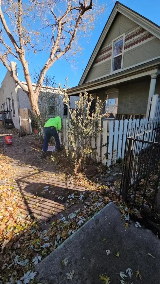 Person in neon green shirt trimming bushes near white picket fence and house. 