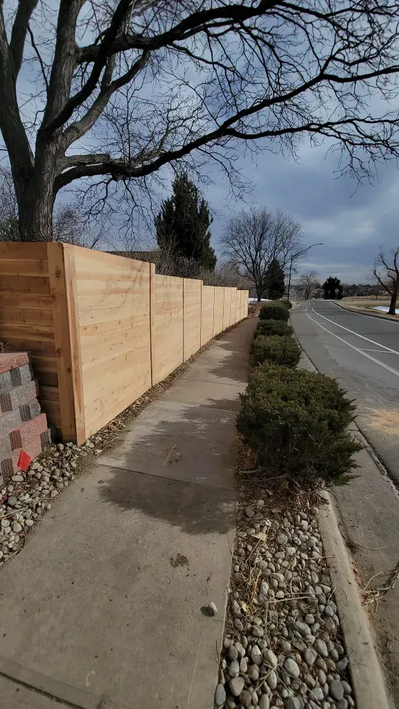 Wooden fence along a sidewalk, with bushes, stones, and a street on a cloudy day.