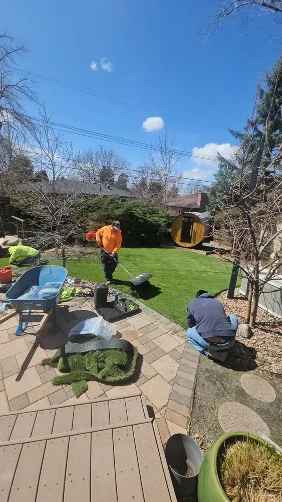 Two people working on a lawn in a backyard. One uses a lawnmower, the other kneels. 