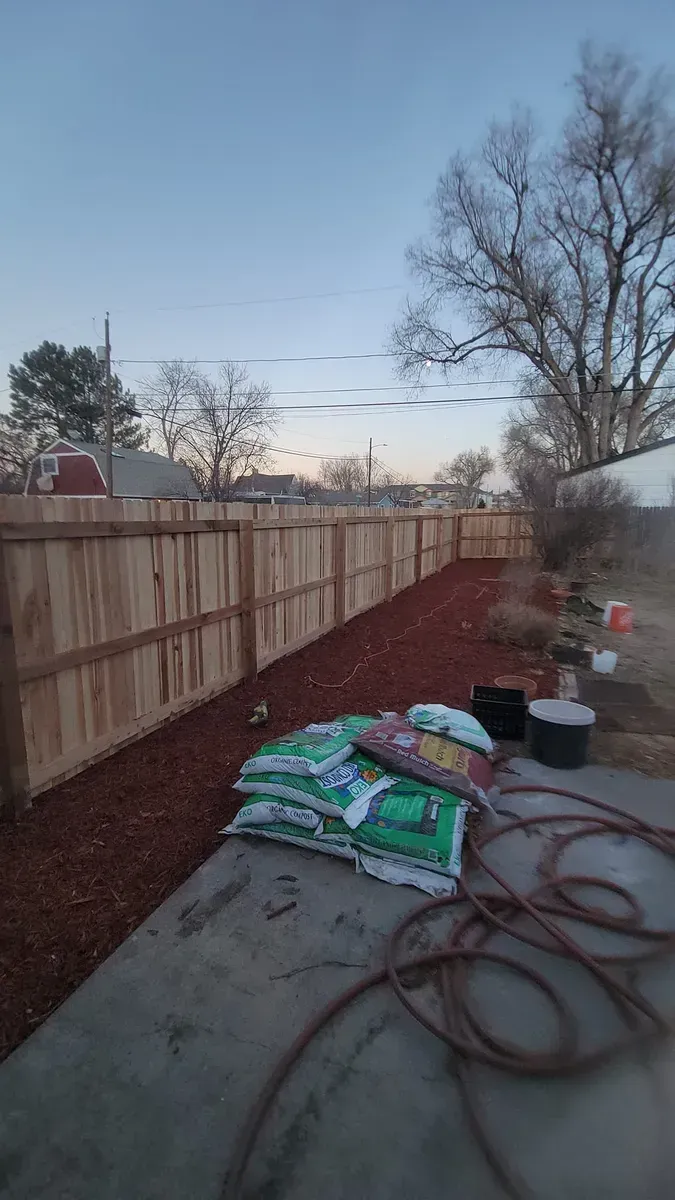 Backyard with a wooden fence, mulch, bags of soil, and a water hose.