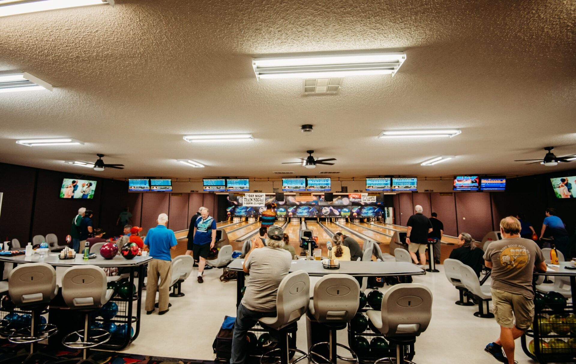 Busy casino gaming room with rows of slot machines and people seated at tables underneath bright monitors